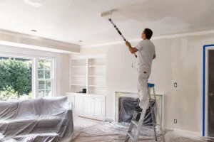 Homme sur escabeau peignant un plafond blanc au rouleau avec bâches de protection au sol