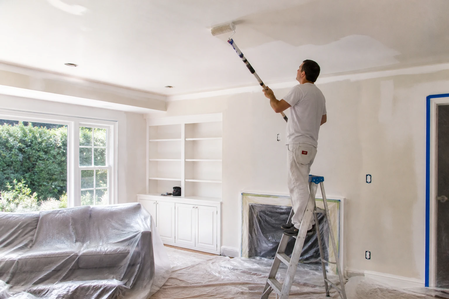 Homme sur escabeau peignant un plafond blanc au rouleau avec bâches de protection au sol